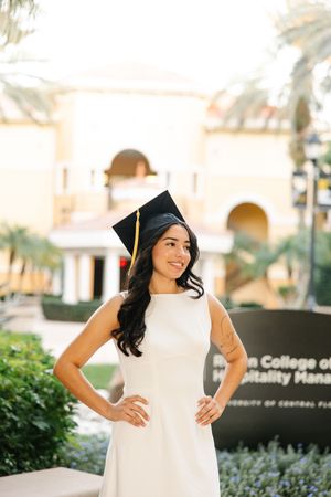 Graduate in white dress poses joyfully outside university building with palm trees.