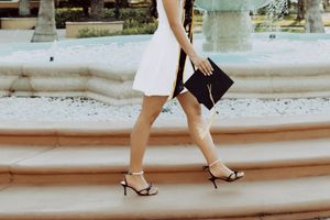 Graduate ascending stone steps in white dress and heels while holding graduation cap.