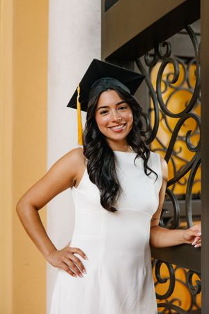 A person in a white sleeveless dress poses elegantly against a yellow wall with ornate iron details.