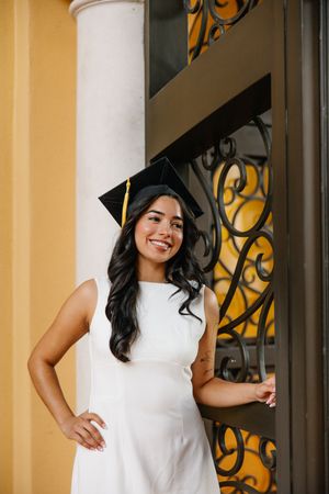 Graduate in white dress poses by ornate wrought iron gate and column.