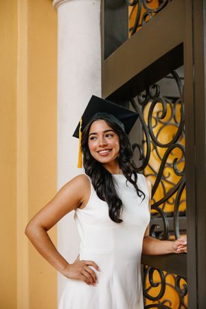 Graduate in cap and gown standing by ornate iron railings and yellow walls.