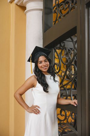 A person in a white dress stands in front of a yellow building with decorative ironwork.