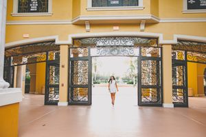 Graduate in white dress stands in ornate yellow archway entrance.