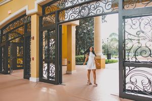 Graduate in white dress stands between decorative iron gates at campus building entrance.
