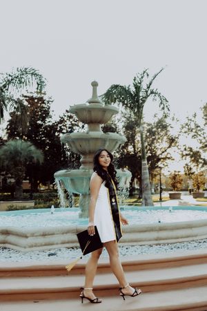 Graduate in white dress walks past decorative water fountain on campus grounds.
