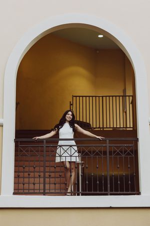 Graduate poses playfully in arched balcony with iron railing.