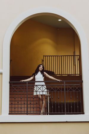 Graduate in white dress poses dramatically on balcony with arched architecture.