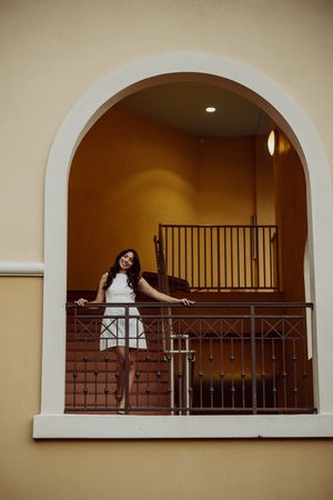 Graduate poses in white dress on an arched balcony with wrought iron railings.