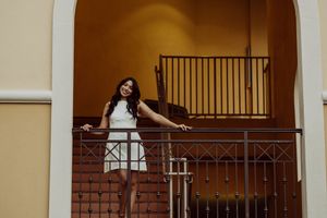 Person in white dress poses on balcony with ornate iron railings and warm lighting.