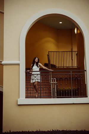Graduate in white dress posing in arched balcony of Spanish colonial style building.