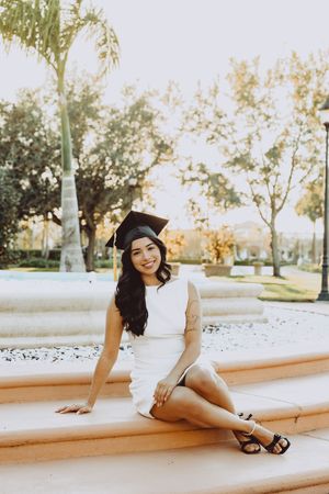 Graduate in white dress and cap poses on stone steps near palm trees at sunset.