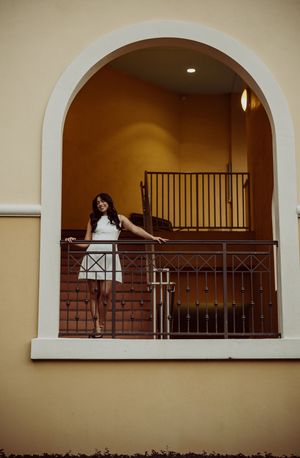 Graduate in white dress poses against railing in arched architectural alcove.