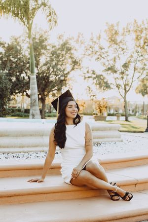 Graduate in sleeveless white dress poses on outdoor stone steps at sunset.