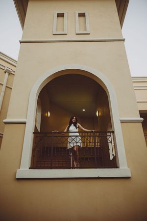 Person in white dress stands on balcony of Mediterranean-style building with arched windows.