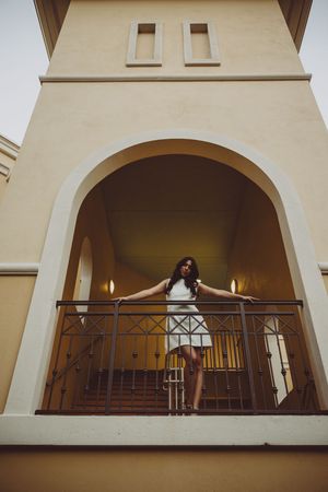A person in a white dress poses dramatically on a balcony with arched architecture in the background.