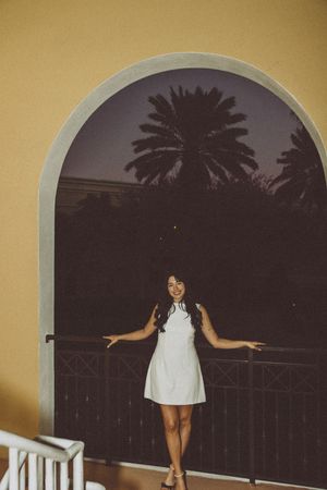 Figure in white dress poses under arched doorway with palm trees silhouetted in background.
