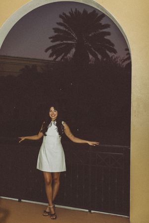 Someone in a white dress stands on a balcony at night with palm trees visible in the background.