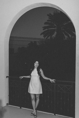 A woman in a white dress poses under an archway at dusk with a palm tree silhouette in the background.