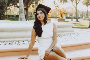 Graduate in white dress sits casually outdoors during sunset photoshoot.