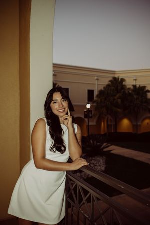 Woman in white dress leaning on balcony railing during sunset with palm trees in background.