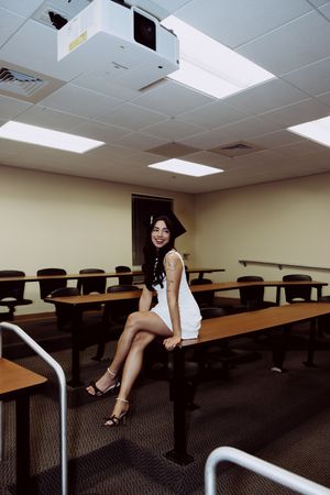 Graduate in white dress and cap poses on classroom desk with projector mounted on ceiling.