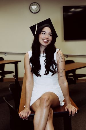 Graduate in white dress sits casually in a classroom wearing a graduation cap and showing tattoos.