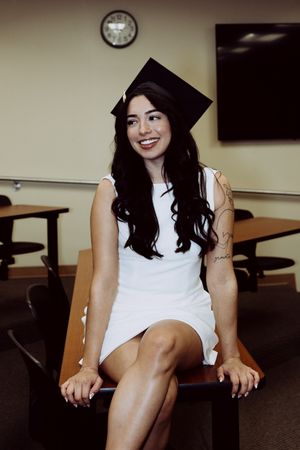 Graduate in academic regalia sitting in classroom setting with clock on wall.