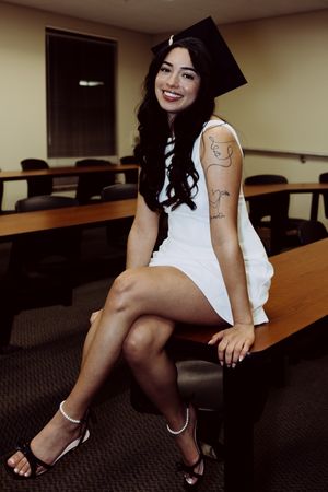 Graduate in white dress and cap sits on classroom desk showing arm tattoo.