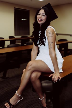 Graduate poses on classroom desk in white dress and cap with visible arm tattoos.