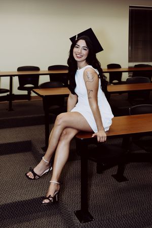 Graduate in cap and white dress sits on classroom desk wearing heels.
