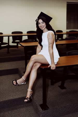Graduate in white dress and mortarboard poses on classroom desk with rows of empty chairs.