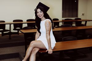 Graduate in white dress poses in modern conference room with dark chairs.