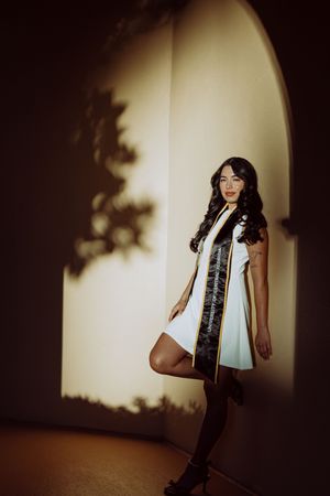 Graduate in white dress and graduation stole poses against wall with dramatic shadow play.