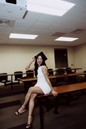 Graduate poses on classroom desk wearing white dress and graduation cap.