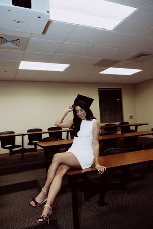 Student in white dress and heels poses playfully in classroom with rows of desks.