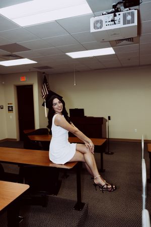 Graduate in a white dress sitting on a bench in a classroom with an American flag in the background.