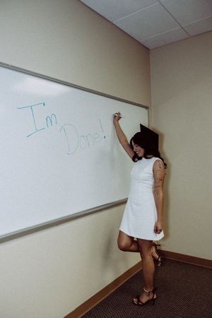 Graduate writes on classroom whiteboard while posing in white dress and cap.