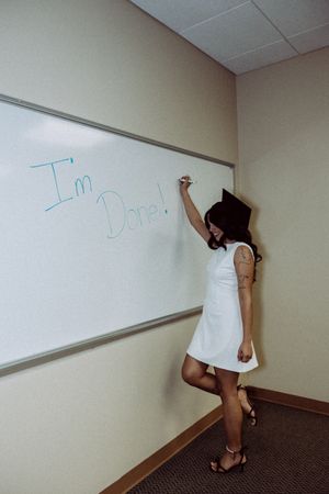 Graduate in white dress writing on whiteboard in classroom setting.