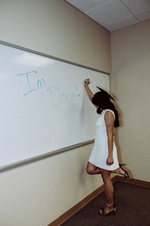 Graduate in white dress writes on whiteboard in classroom.
