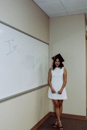 A person wearing a graduation cap and white dress poses in front of a classroom whiteboard.