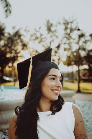 Graduate in white dress and cap looks to side with joyful expression during sunset photo session.