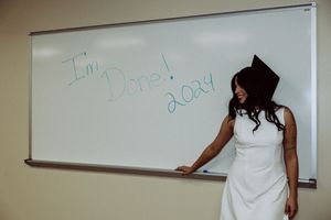 Graduate in white dress writes on whiteboard in classroom setting.