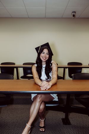 Graduate sits at classroom table in white dress and graduation cap.