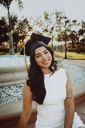 Graduate wearing cap and gown sits by fountain during golden hour sunset.