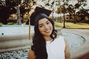 Graduate in cap and gown smiles brightly near fountain during outdoor graduation photoshoot.