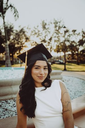 Graduate in cap and white dress smiles while standing near outdoor fountain.