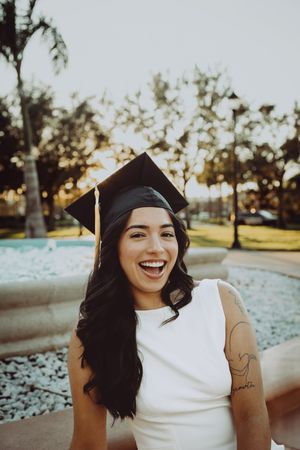 Graduate in white dress and mortarboard laughs joyfully during outdoor graduation photos by fountain.