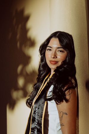 Graduate in sash poses against wall with dramatic shadow play.