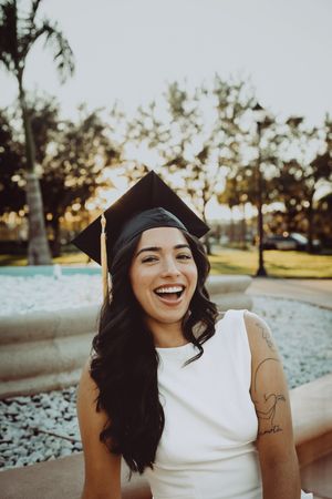 Joyful graduate laughing in cap and white dress during outdoor photoshoot.