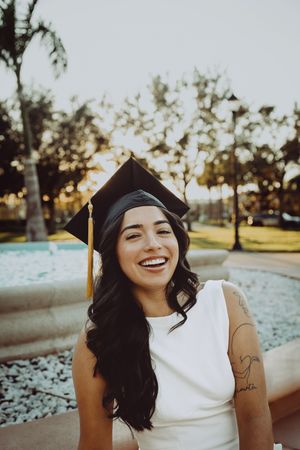 Graduate in academic regalia smiles while sitting outdoors during golden hour photography session.